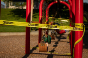 Empty saddle style swings at a playground covered in yellow caution tape written in english and spanish.