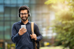 Smiling young indian man wearing wireless headphones holding phone having video call. Eastern businessman using smartphone, listening music in app tech on cellphone watching videos online, copy space