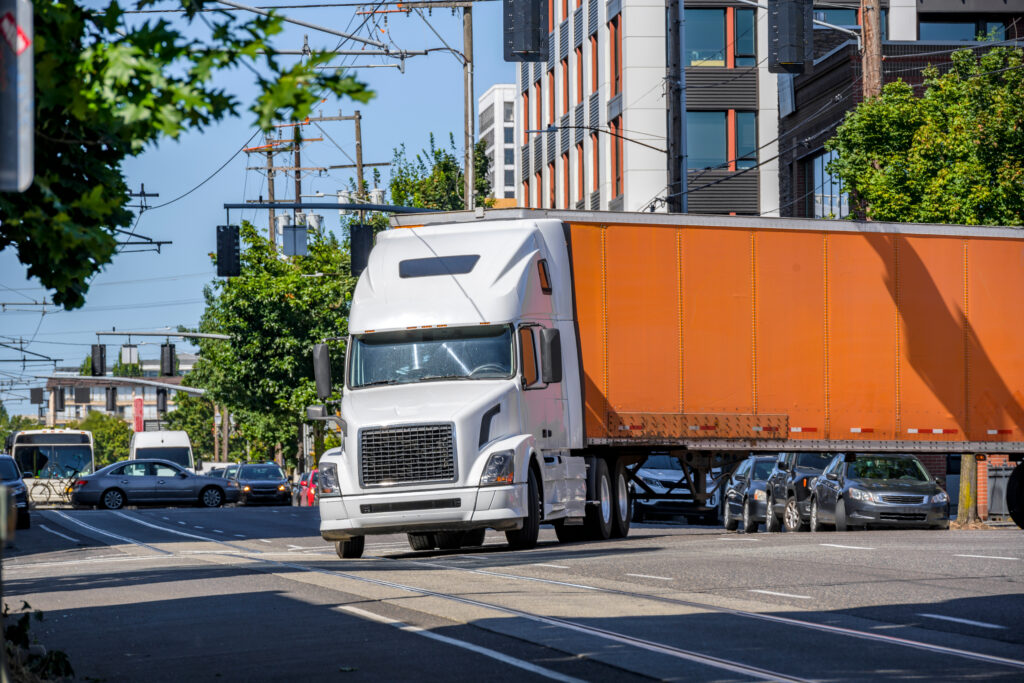 White classic industrial diesel big rig semi truck tractor transporting commercial cargo in orange dry van semi trailer turning on the crossroad street intersection with busy city traffic