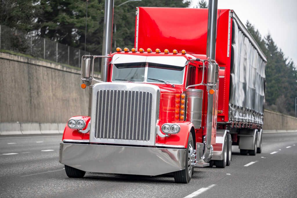 Classic big rig red semi truck tractor with chrome high exhaust pipes and truck driver sleeping compartment transporting cargo in covered semi trailer running on the road with wall on the side