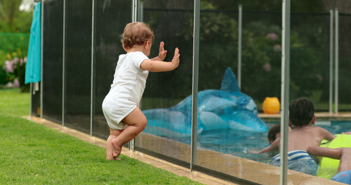 Cute candid moment of baby leaning on pool fence wanting to interact with kids inside swimming pool