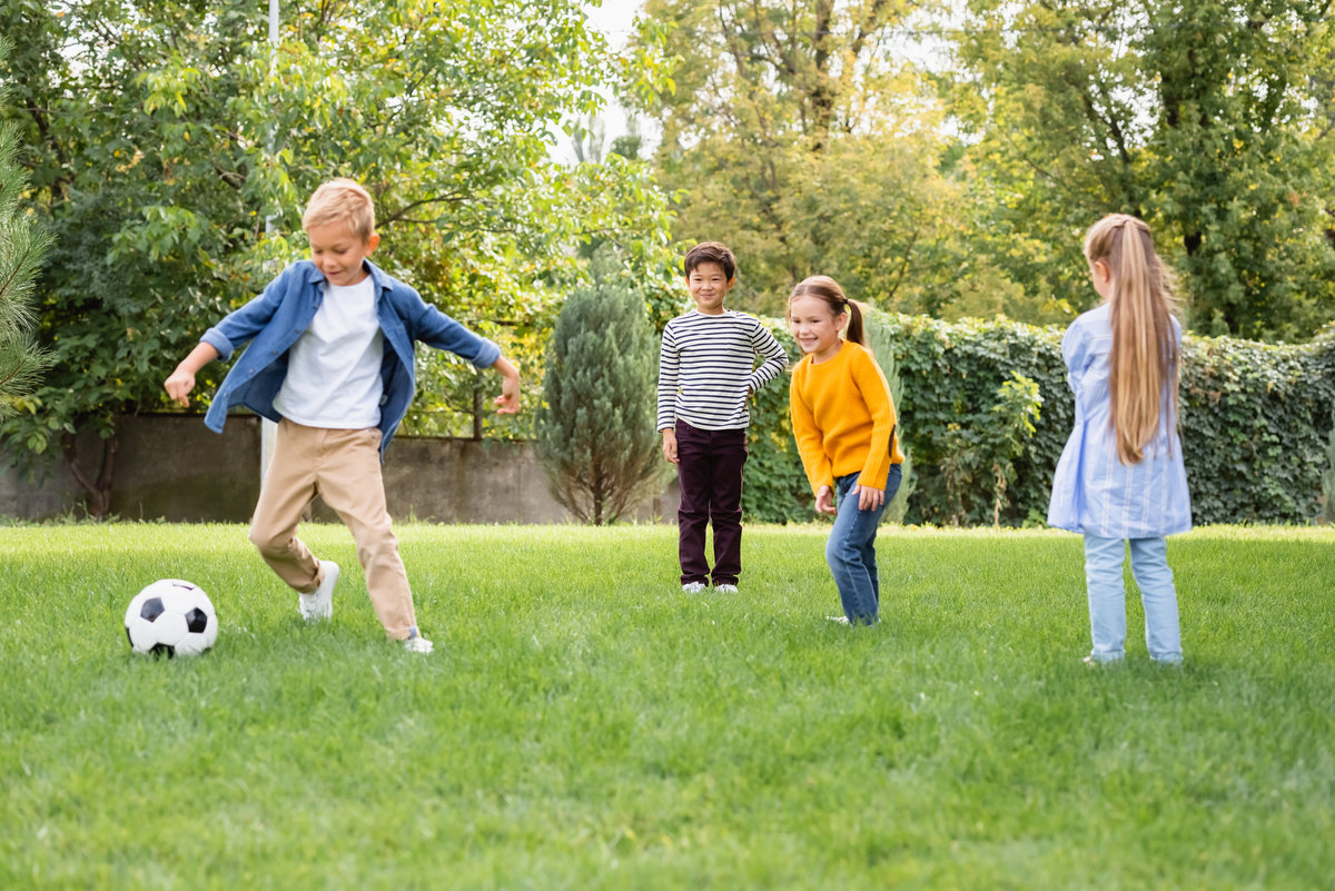 Positive multiethnic children standing near friend playing football in park